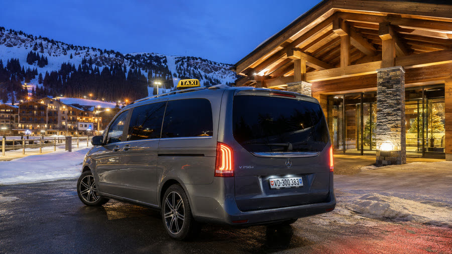 Taxi Mercedes Classe V devant un hôtel de montagne à la tombée du jour, station de ski illuminée — dépose à l'hôtel comprise