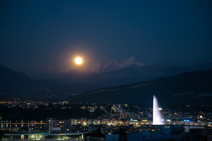 Le Jet d’eau de Genève illuminé la nuit, la lune au-dessus du Mont-Blanc et la ville allumée — taxi Lausanne–Genève 24h/24